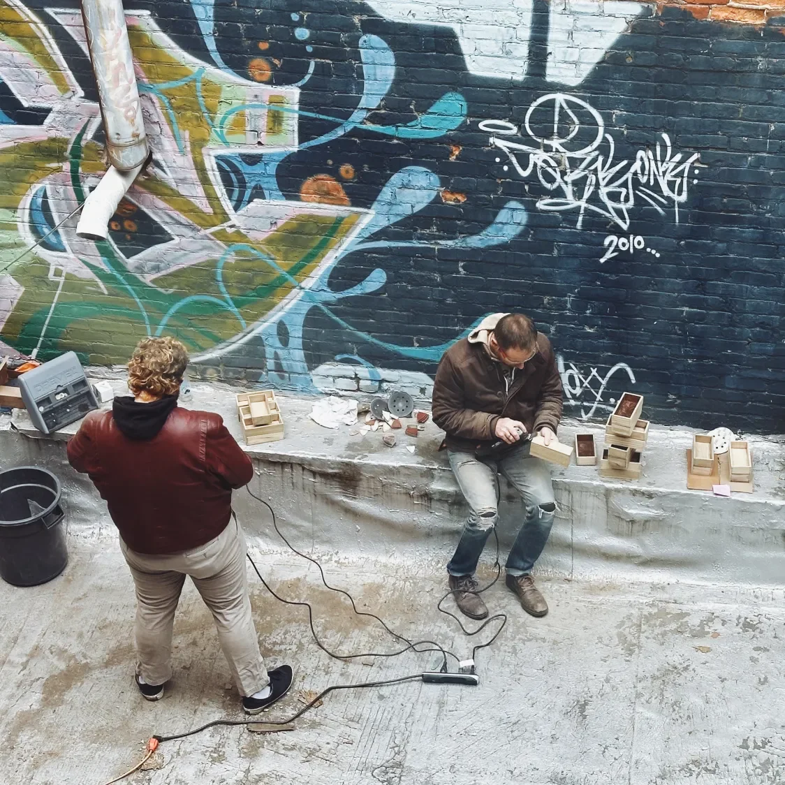 Chris Rinehart and Gabriel Cuddy sanding wood boxes against a graffitied wall