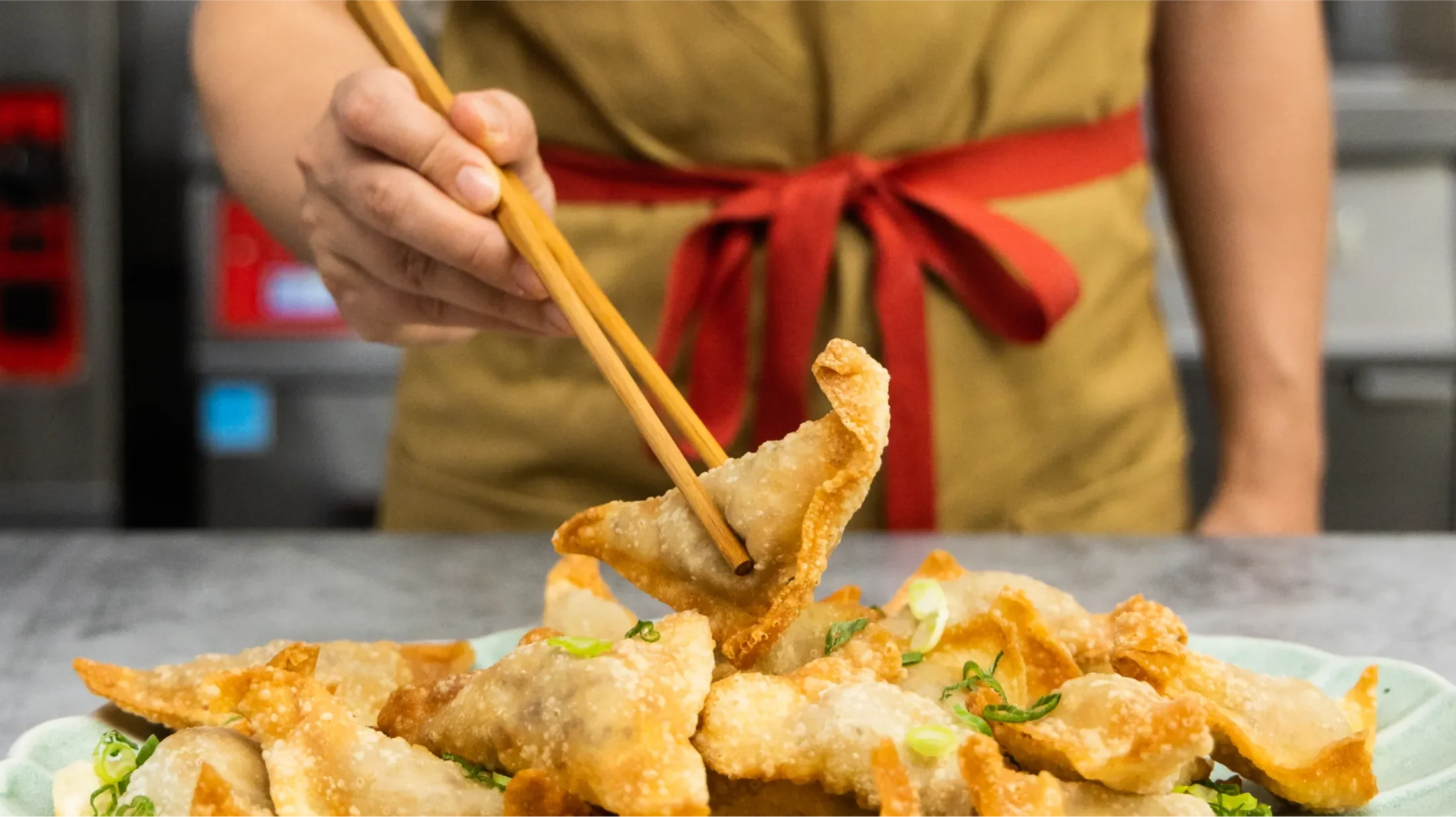 Man with a red sash tied around his waist holding a fried wonton with chopsticks over a full plate